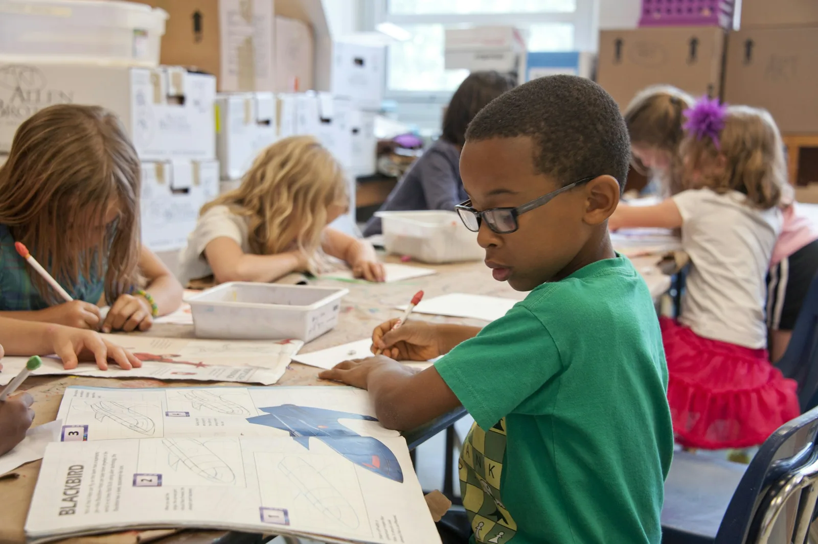 A pupil drawing at a classroom desk, with classmates nearby.