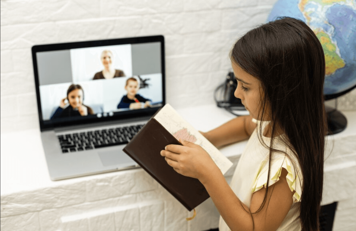 A child holding a book beside a laptop video call.