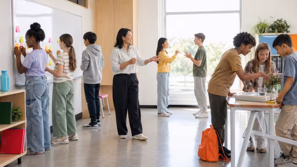 Real K-12 classroom mid-activity: diverse students at whiteboard with sticky notes, two role-playing, group at hands-on science table; teacher gesturing in middle