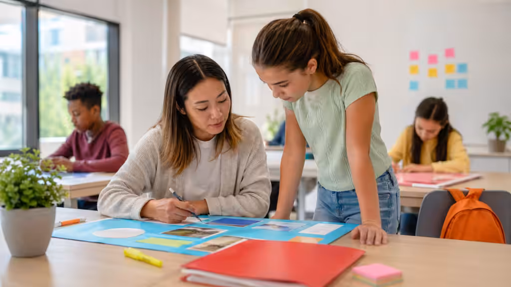 Teacher reviewing a student's poster project at a desk in a modern bright classroom; the student leans in; other students working in soft-focus background