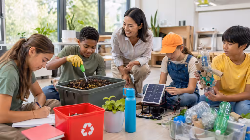 Una docente y varios estudiantes exploran materiales, plantas y un pequeño panel solar en un aula.