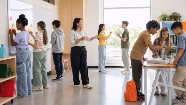 Escena real en un aula de primaria y secundaria durante una actividad: alumnos de diversa procedencia frente a la pizarra con notas adhesivas, dos realizando un juego de roles, un grupo en la mesa de ciencias con actividades prácticas; el profesor gesticulando en el centro
